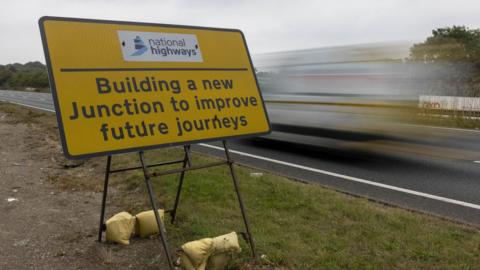 A yellow sign beside a road with the National Highways logo at the top, with the words 'building a new junction to improve future journeys' below. There is a blurred vehicle on the road to the reside of the sign