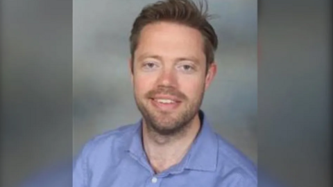 A man in a blue shirt is looking directly at the camera and smiling