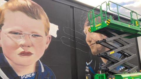 A view of the mural currently being painted. A young Ed Sheeran with ginger hair and glasses can be seen while an older version of himself is painted next to it and hidden behind a green scissor lift. 