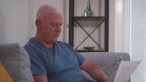 Paul Harrison has short grey hair and is wearing a blue t-shirt. He is sitting on a grey sofa in his living room and reading a letter.