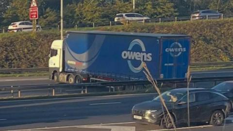 A blue lorry with the word Owens on the side is stationary across the lanes of the motorway carriageway near a slip road as cars drive past on other side of motorway.