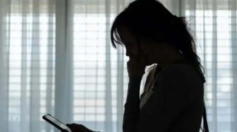 A woman stands by a window and looks at a phone with her hands resting on her chin. She has long hair but is pictured in silhouette.