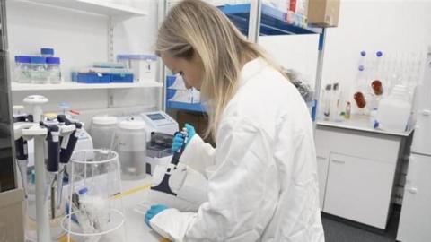 A scientist in a white lab coat working on a vaccine. She has blonde hair and is in a lab surrounded by equipment.