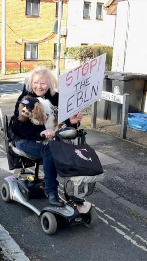 A woman on a mobility scooter, holding her dog and a sign saying Stop the EBLN