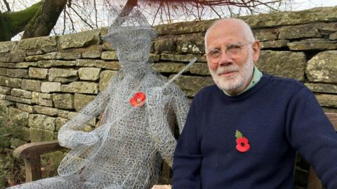 Sam Simson sat on a bench next to a life size human chicken wire model holding a banjo.