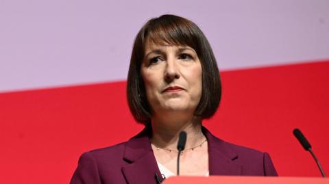 Rachel Reeves with a thoughtful look on her face standing behind a podium during a speech. She is dressed in a purple jacket. 