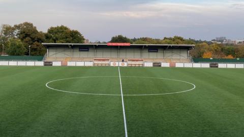 harlow Town football pitch shows the central bit of the grassed area with a circle outlined in white being dissected by a white line. A covered seating stand is in the background with a low fence before it with advertising boards on. 