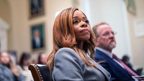 A woman with reddish-brown hair looks ahead in a grey suit. She is sitting in a brown chair. In the background, other people sitting in chairs can be seen, but are blurry.