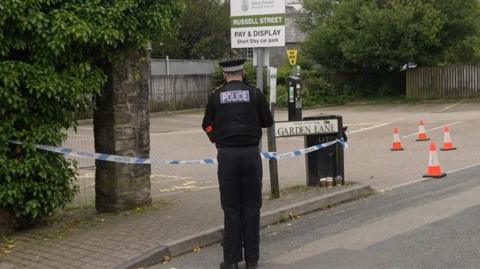 A police officer is seen standing next to a car park with his back to the camera. There are bollards around the area and police tape. 