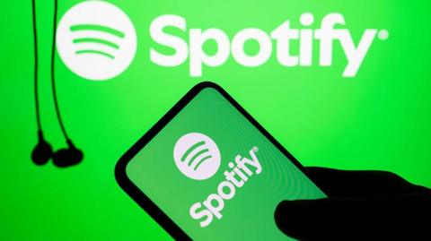The silhouette of a hand holds a phone with the green and white Spotify logo on it. In the background hang some black wired earbuds.