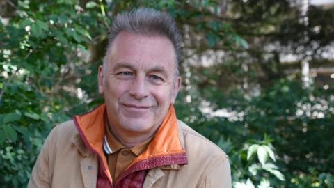 Chris Packham, sitting outside in front of green foliage. He is looking at the camera and smiling and is wearing an orange T-shirt with a red coat and a brown coat over the top.