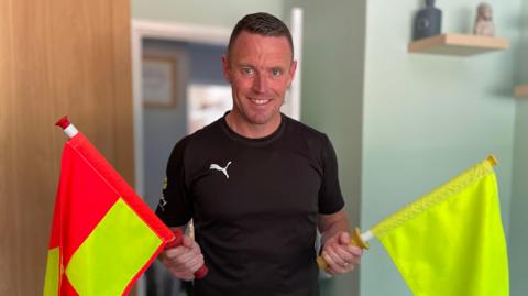 Stuart Burt in a black Puma refereeing kit. In one hand he is holding a bright yellow and orange chequered flag, while in the other he is holding a plain yellow flag.