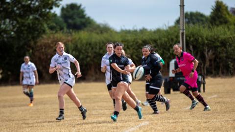 A women's rugby sevens game taking place on a pitch. A player dressed in a dark blue kit is passing the ball to her left as other players run alongside and watch her. 