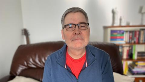 John Gidman is wearing a red t shirt and blue quarter zip jumper, he is sitting on a sofa in front of a bookcase, a guitar is propped up in the corner of the room behind him.