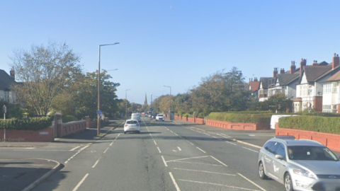 Cars driving along a road, with houses to either side. There is a junction with road turning off to the left and right in the centre of the image, and a church spire in the distance.