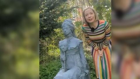 Lucy Worsley standing with statue of Princess Caraboo in a burial ground in Bristol. Lucy Worsley is wearing a rainbow stripy dress and the statue is a grey colour.