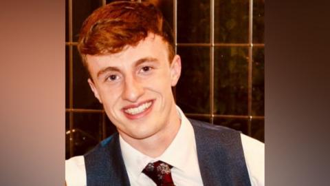 A man with auburn hair in a waistcoast and white shirt with a claret tie