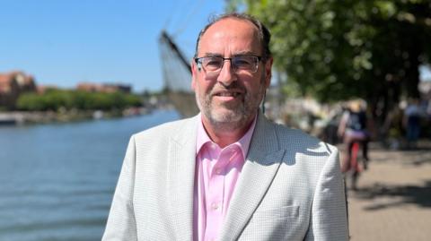 Councillor Tony Dyer. He is wearing a light grey suit with a pink shirt. He is wearing glasses and is standing by Bristol harbour.