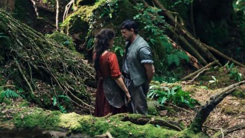 A still of the film Hamnet, showing Jessie Buckley as Agnes and Paul Mescal as William Shakespeare. The pair stand in period dress very close together in the middle of a forest.
