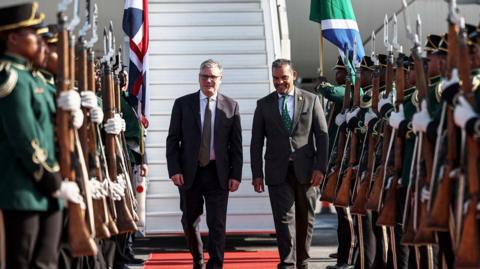 Keir Starmer lands in South Africa and is greeted by a South African official . He walks past people in uniform holding rifles