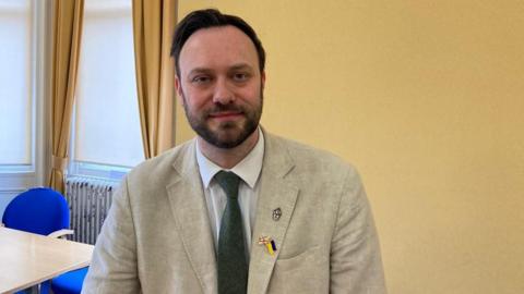 Middlesbrough councillor Luke Henman. He has short dark hair and a trimmed beard. He is wearing a cream jacket, white shirt and green tie. He has three pins on his lapel, including a union jack pin and a Ukraine flag. He is looking at the camera and smiling. He is in a room with yellow walls and yellow curtains on a window. Behind him there is a table with blue chairs.