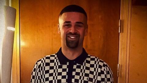 Tariq Evans, who has a shaved head and eyebrow, has a dark beard and moustache and is wearing a black and white check polo shirt