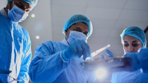 Three surgeons wearing face masks, gloves and blue scrubs look down at some surgical tools in preparation for an operation.