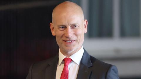 A bald man in a dark suit, white shirt and red tie.