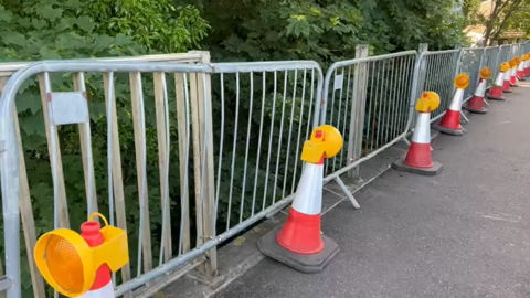 A bridge with temporary metal railings placed along the edge. Orange and white traffic cones are lined in front of the barriers. 