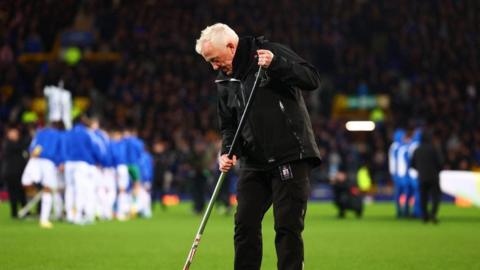 Bob Lennon, in his 60s, stands on a football pitch holding a garden hoe against the green turf. In the background a group of players wearing blue training tops are stretching, while the stands are full of spectators. Lennon has white hair and is wearing a black coat, black jeans and black shoes. 