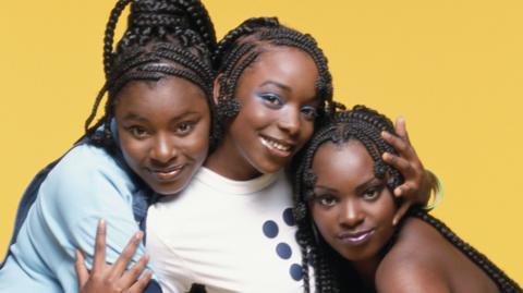 Sisters Cleo, Yonah, and Zainam Higgins of the group Cleopatra look into the camera as they hug each other. They all have black braided hair. Yonah and Zainam, wearing blue tops, stand either side of Cleo, who is wearing a white T-shirt with a black dot design on the front. The background is a yellow wall