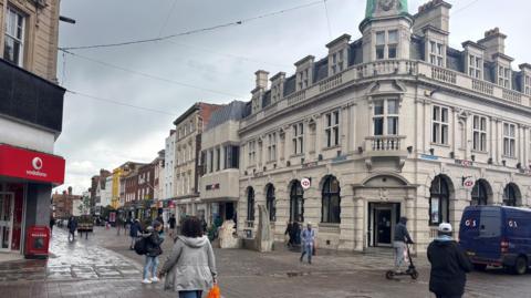 A rainy day at an historic city centre crossroads. People are crossing over on foot and one person is on an e-scooter. A Vodafone shop can be seen on one side and the HSBC on the other.