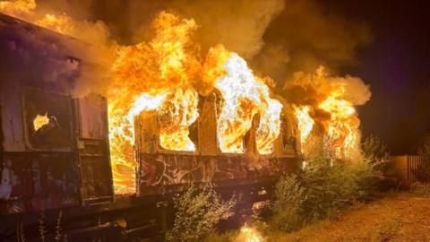 A train carriage with flames bursting out of the windows. There are bushes growing near the carriage, and fencing in the distance.