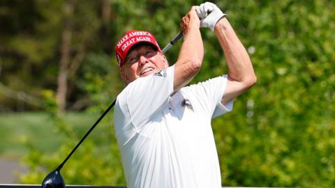 Donald Trump playing golf, mid-swing, frozen in a poised follow-through on the golf course . He’s dressed in a white polo shirt and gloves, and wears a red cap emblazoned with the words “MAKE AMERICA GREAT AGAIN.