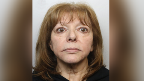 A woman with shoulder-length dark blonde hair is looking at the camera in a police custody shot. There is a Greater Manchester Police logo in the top left hand corner.