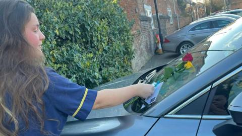 A woman is standing on the left and is wearing a blue tunic. She has long brown hair and is putting flowers and a white card on the front window screen on a silver car.