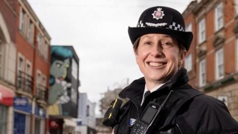 Rachel Nolan wearing black uniform with a blue police badge and hat. She is standing on a busy Chelmsford High Street scene and is smiling at the camera and her hands are behind her back