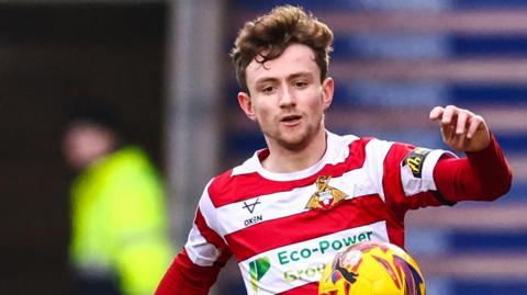 James Maxwell is wearing a red and white hooped Doncaster shirt and is trying to control the ball as he runs forward.