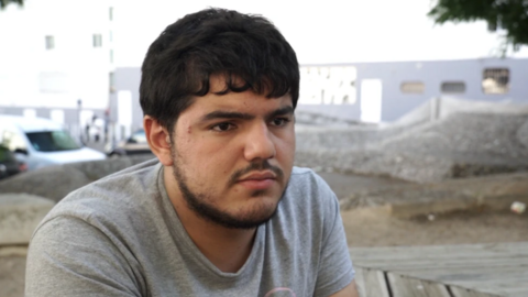 A young man with a beard wears a grey T-shirt and stares out with a backdrop of Marseille