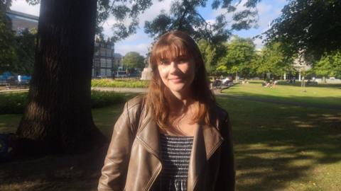 Amala Parry with long brown hair, wearing a striped top and brown leather jacket, looks at the camera as she stands in front of a tree on grass. 