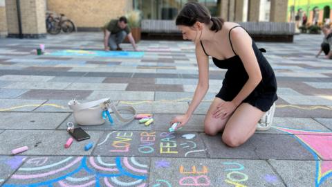 A person kneels on a tiled outdoor surface, drawing colorful chalk art that includes the phrases 'LOVE OVER FEAR' and 'BE YOU'. Chalk pieces, a white handbag, and a smartphone are scattered nearby. Another person is also drawing in the background, with buildings and benches visible in the urban setting.