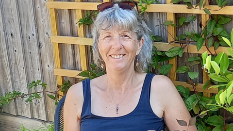 Jan sat in front of a garden fence. She has short grey hair and wears a navy vest, a necklace and brown sunglasses on her head. She is smiling.