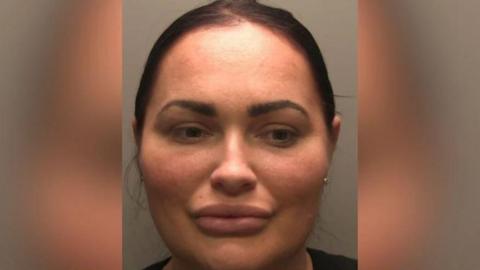 A police custody picture of Natasha Allarakhia. She has black hair tied back and is looking to the side. The wall behind her is grey. 