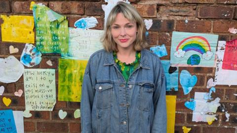 Jodi Ann Bickley standing in front of a wall displaying colourful and uplifting messages. She is wearing a denim jacket and smiling. One of the messages reads: "How lucky am I to exist on this massive planet right next to you? Very lucky indeed." 