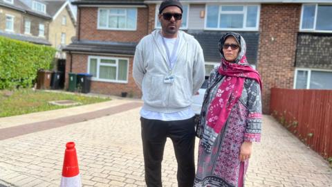 A man and woman standing in a driveway in front of a house. There is a traffic cone next to them. They look like a mother and son. The man wears a light grey zip-up top and black trousers. The woman wears a traditional South Asian shalwar-kameez outfit.