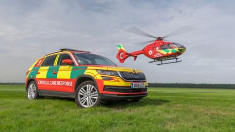 A car and a helicopter belonging to Thames Valley Air Ambulance photographed on a cloudy day. The parked car has a sign on the side that reads "Critical Care Response". The helicopter is in the air.