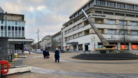 A general view of Plymouth City Centre. The sundial is on the right and there's people stood nearby. The toilet blocks are to the left. 