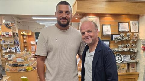 Ben Miller, a man with short dark hair, a beard and a beige shirt, stands next to actor Toby Jones, who is wearing a white shirt and dark blue jacket. They are both standing in a pottery gift shop.
