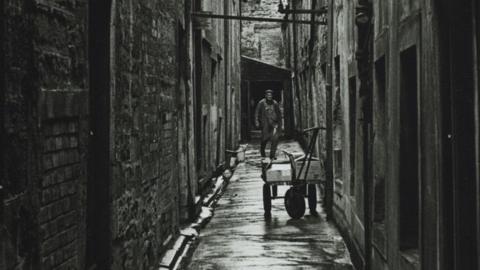 A black and white photo of a narrow passageway with a workman and a cart 
