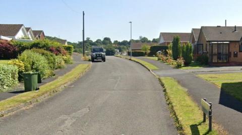 A Google image shows one vehicle parked on a road with bungalows either side and green verges. Various plants and shrubs can be seen in the gardens. There are trees in the distance.